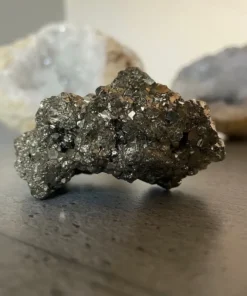 Pyrit surový kameň S. nepravidelný kus minerálu. Celkový tvar je drsný a hrboľatý. Okraje sú nepravidelné, miestami zaoblené, miestami ostrejšie. Pripomína to zhluk menších kamienkov spojených do jedného celku. Farba a lesk: Kameň má výraznú striebristo-sivú, kovovú farbu. Jeho povrch intenzívne leskne a odráža svetlo. Tento lesk nie je hladký a jednotný ako leštený kov, ale je rozptýlený vďaka nespočetnému množstvu drobných, trblietavých plôšok. Predstavte si, ako sa leskne rozdrvený kov alebo tisíce malých, jemných zrkadielok spojených do jedného celku. Farba je konzistentne striebristá po celom povrchu. Štruktúra a povrch: Ide o tzv. drúzu, čo znamená, že povrch nie je hladký, ale je pokrytý nespočetnými drobnými kryštálmi, ktoré rastú vedľa seba. Vďaka tomu je povrch extrémne drsný, hrubý a veľmi štruktúrovaný. Je plný malých výstupkov, zrniečok a preliačin. Môžete si to predstaviť ako veľmi hrubý brúsny papier s kovovým leskom, alebo ako špongiu, ktorá je namiesto pórov plná malých, trblietavých kryštálikov. Niektoré miesta môžu mať mierne väčšie, zreteľnejšie kryštálové plôšky, ktoré sa lesknú intenzívnejšie.