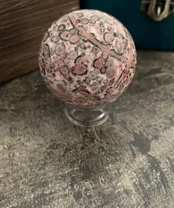Jaspis pink leopard guľa. Hladký, leštený kameň guľového tvaru s dominantnými farbami rôznych odtieňov ružovej — od jemnej púdrovej až po sýtu staroružovú. Doplnený je kontrastnými oblasťami sýtej čiernej, tmavosivej až bridlicovej, ako aj svetlosivej a béžovej s teplým pieskovým nádychom. Farby sa prelínajú v prirodzených prechodoch a vytvárajú vrstvené škvrny a prstence. Povrch pokrývajú výrazné kruhové a oválne vzory pripomínajúce leopardiu srsť, pričom tmavé stredy sú často orámované ružovými alebo svetlými prstencom, čo vytvára dojem organického, prírodou formovaného dizajnu.