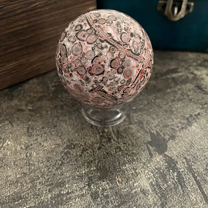 Jaspis pink leopard guľa. Hladký, leštený kameň guľového tvaru s dominantnými farbami rôznych odtieňov ružovej — od jemnej púdrovej až po sýtu staroružovú. Doplnený je kontrastnými oblasťami sýtej čiernej, tmavosivej až bridlicovej, ako aj svetlosivej a béžovej s teplým pieskovým nádychom. Farby sa prelínajú v prirodzených prechodoch a vytvárajú vrstvené škvrny a prstence. Povrch pokrývajú výrazné kruhové a oválne vzory pripomínajúce leopardiu srsť, pričom tmavé stredy sú často orámované ružovými alebo svetlými prstencom, čo vytvára dojem organického, prírodou formovaného dizajnu.